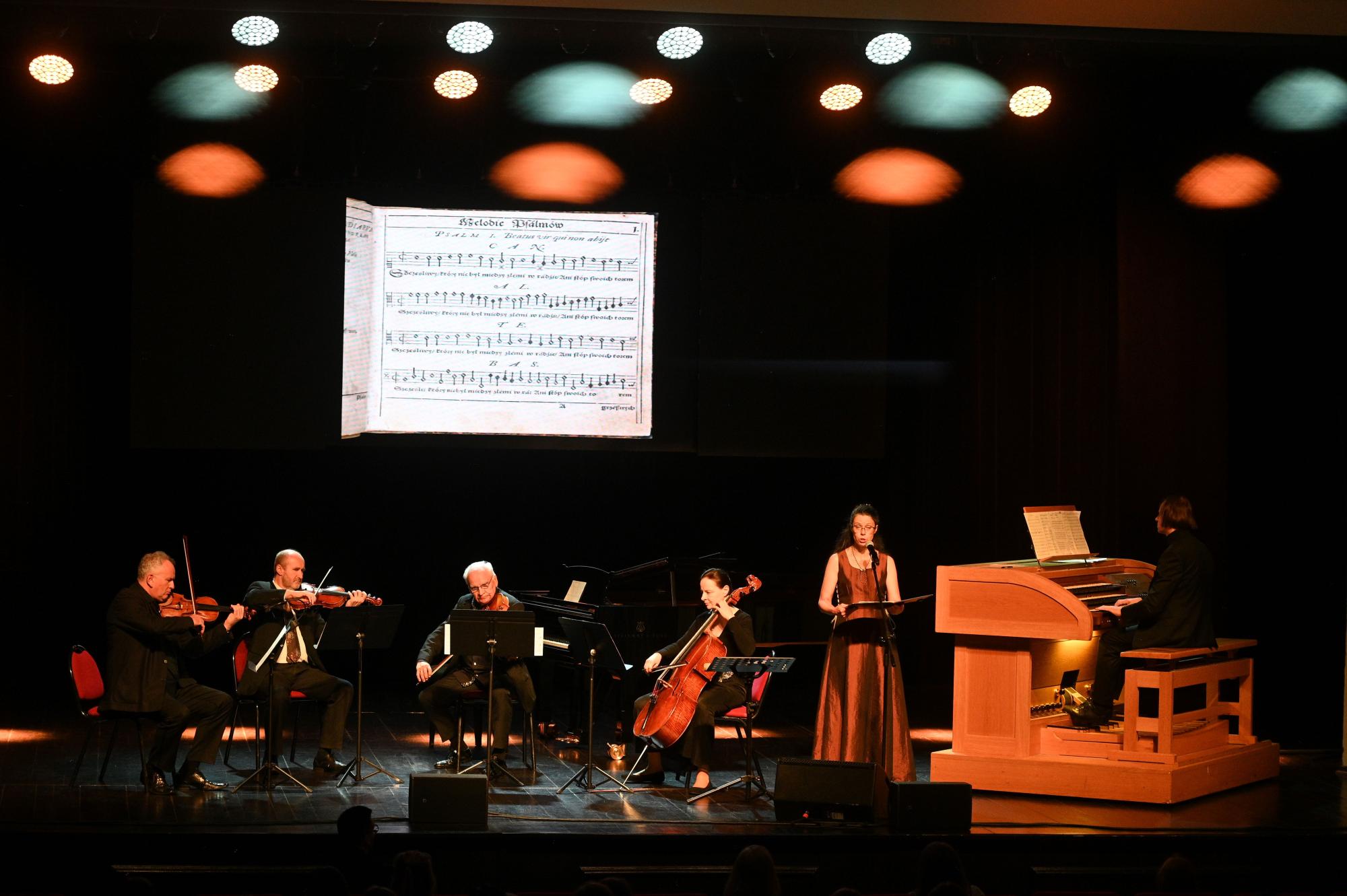 Czwórka muzyków gra na skrzypcach, altówce, wiolonczeli, obok stoi śpiewaczka trzymająca nuty, obok organista przed klawiaturą instrumentu