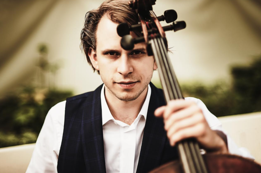 Wiolonczelista Marcin Zdunik trzyma w lewej ręce instrument, ubrany jest w białą koszulę i czarną kamizelkę