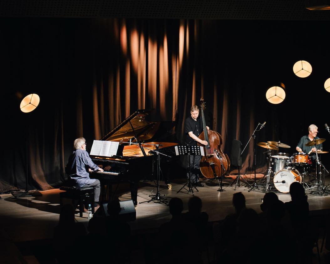 Trio Andrzeja Jagodzińskiego, pianista siedzi przy czarnym fortepianie, kontrabasista stoi z instrumentem, perkusista siedzi za składem instrumentów