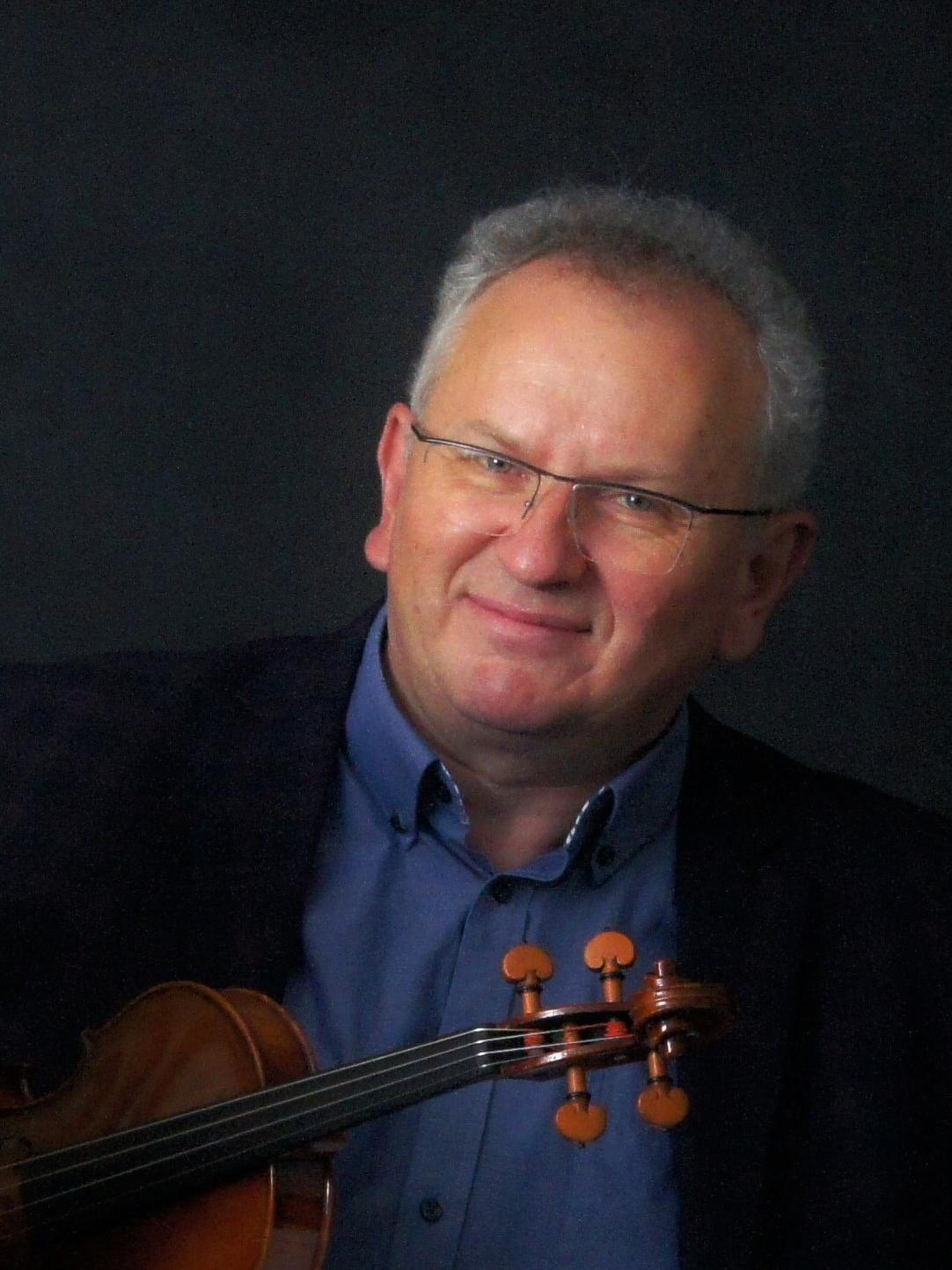 Skrzypek Adam Wagner pozuje ze skrzypcami, jest ubrany w niebieską koszulę i czarną marynarkę, nosi okulary