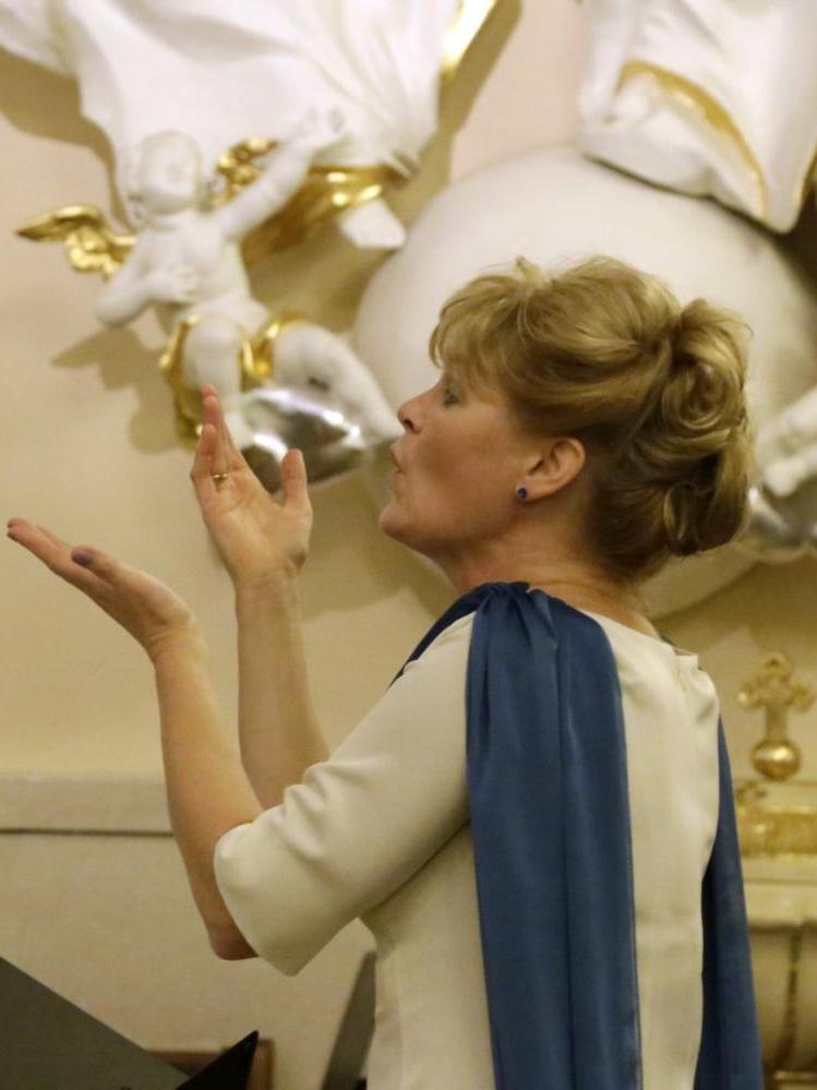 Dyrygentka chóralna Beata Borowska dyryguje utrworem muzycznym, jest ubrana w jasną sukienkę i niebieski szal