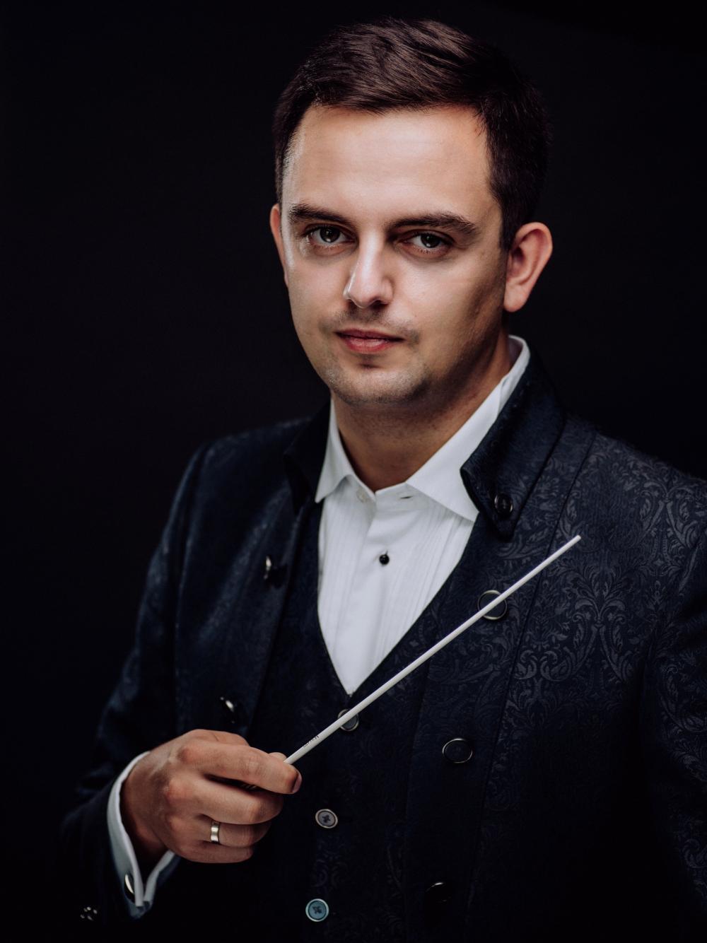Młody dyrygent Maciej Tomasiewicz trzyma w oprawej ręce batutę i ubrany jest w szarą ozdobną marybarkę i białą koszulę