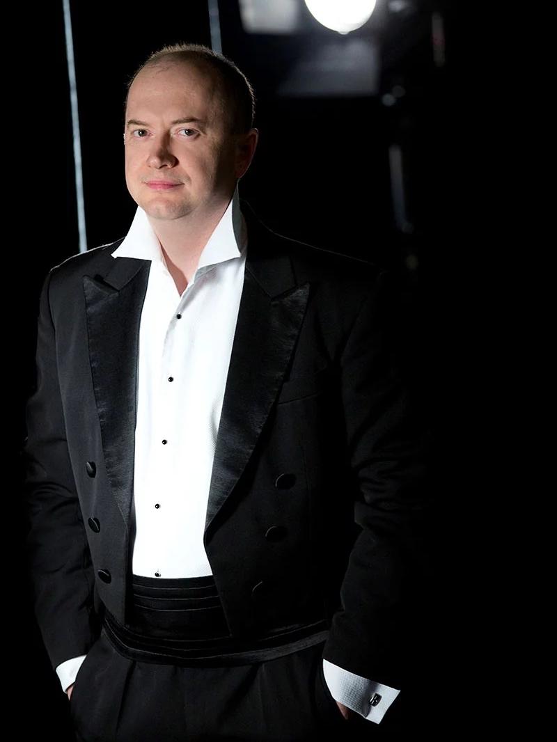 Dyrygent Michał Klauza ubrany jest w białą koszulę i czarną marynarkę