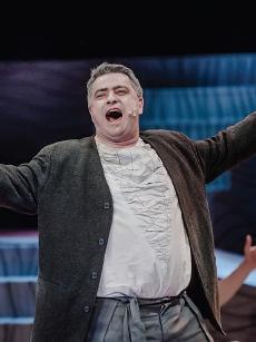 Tomasz Lorek jest śpiewakiem w średnim wieku, występuje w białej koszuli i brązowym surducie, ręce ma szeroko rozłożone